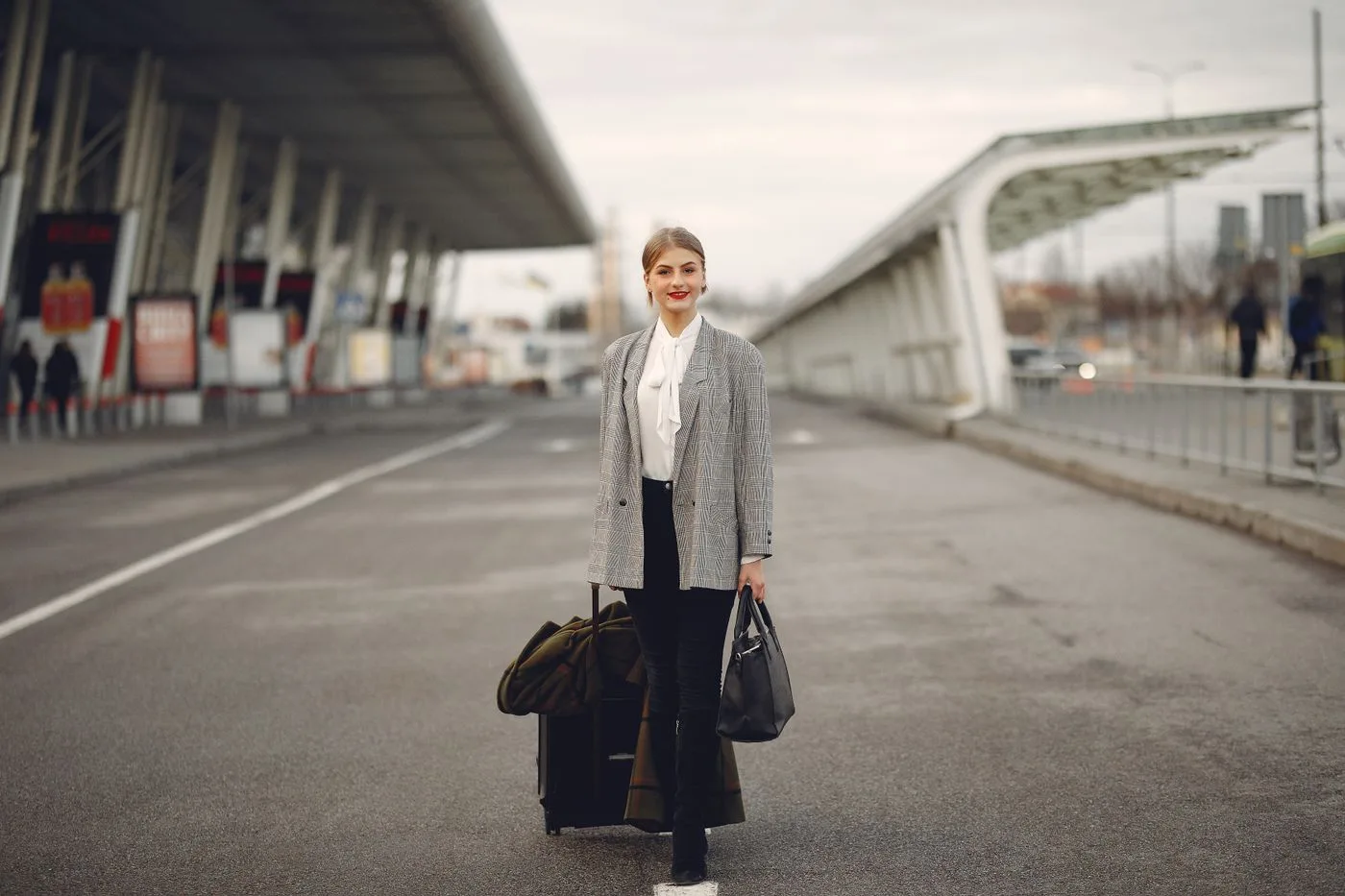 A woman in a gray coat walking with luggage outside a terminal.