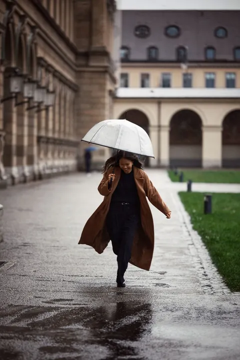 A woman in a brown coat walks with an umbrella along a wet city street.